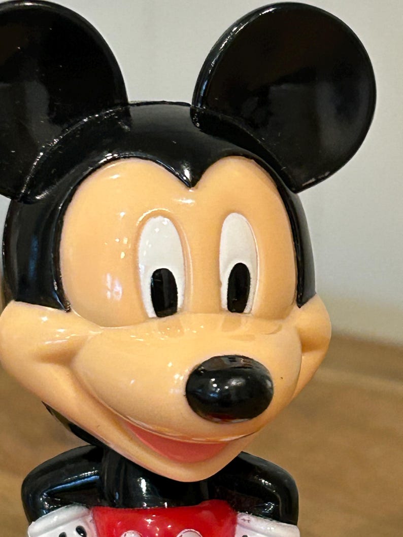 May include: A close-up of a Mickey Mouse figurine. The figurine has a black head and ears, a tan face, and a black nose. The figurine is wearing a black jacket with a red bow tie.