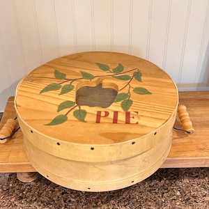 Può includere: Un contenitore rotondo in legno per torte con un coperchio con un ritaglio a forma di mela e la parola "PIE" in rosso. Il coperchio è decorato con foglie verdi. Il contenitore ha due manici in legno e una rete.