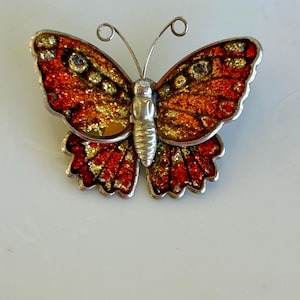 May include: A butterfly brooch with a silver-tone metal frame. The wings are a mix of red, orange, and gold, with a glittery effect. The body is silver, and the antennae have curled tips. A decorative accessory.