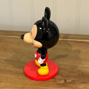 May include: A Mickey Mouse figurine with a black head and ears, a tan face, and a red and white outfit. The figurine stands on a red circular base. The figure is a classic Disney character.
