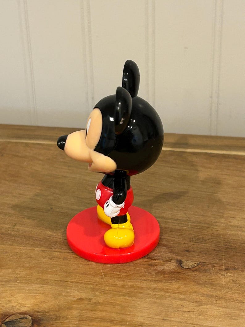May include: A Mickey Mouse figurine with a black head and ears, a tan face, and a red and white outfit. The figurine stands on a red circular base. The figure is a classic Disney character.