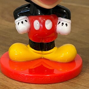 May include: A Mickey Mouse figurine with a red base and shorts, yellow shoes, a black jacket, and white gloves. The figurine has a smiling face and is a collectible item.