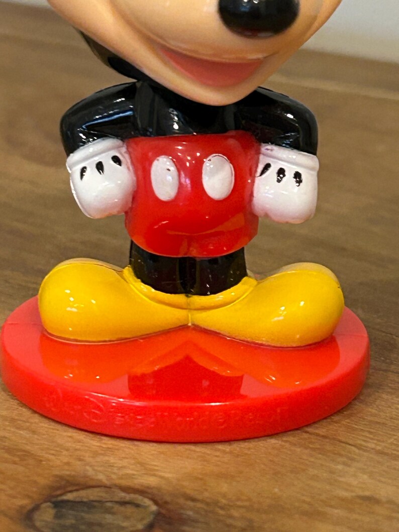 May include: A Mickey Mouse figurine with a red base and shorts, yellow shoes, a black jacket, and white gloves. The figurine has a smiling face and is a collectible item.