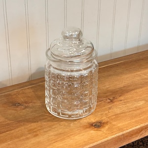 May include: A clear glass canister featuring a textured grid pattern on the exterior walls. The cylindrical container includes a matching removable glass lid with a rounded knob handle at the top, providing a secure cover for the storage vessel.
