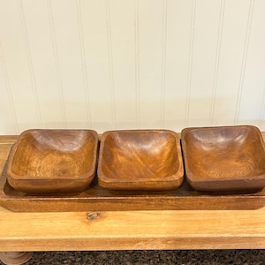 Pode incluir: Uma bandeja de madeira com três tigelas quadradas. As tigelas e a bandeja são feitas de madeira castanha clara. As tigelas são colocadas na bandeja, que tem uma borda ligeiramente elevada. A bandeja está sobre uma superfície de madeira.