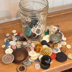 May include: A clear glass mason jar with a diamond-patterned texture filled with a variety of assorted sewing buttons in different sizes, shapes, and colors, including brown, white, black, gold, red, and blue, scattered around the base.