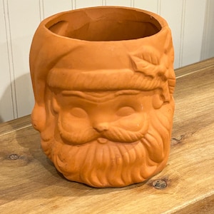 May include: A terracotta Santa Claus head-shaped planter. The planter has a detailed face with a beard, mustache, and hat. The open top is designed for planting flowers or other decorative items. The planter is a warm orange color.
