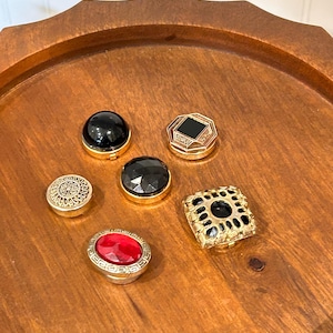 May include: Six decorative buttons in a variety of designs and colours. The buttons feature black, red, and clear gemstones set in gold-toned metal. The buttons are arranged on a wooden tray.