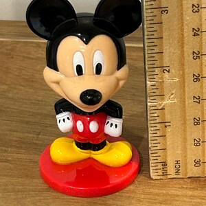 May include: A Mickey Mouse figurine with black ears, a tan face, and a red and white outfit. The figurine is standing on a red base. A wooden ruler is next to the figurine, showing measurements in inches.