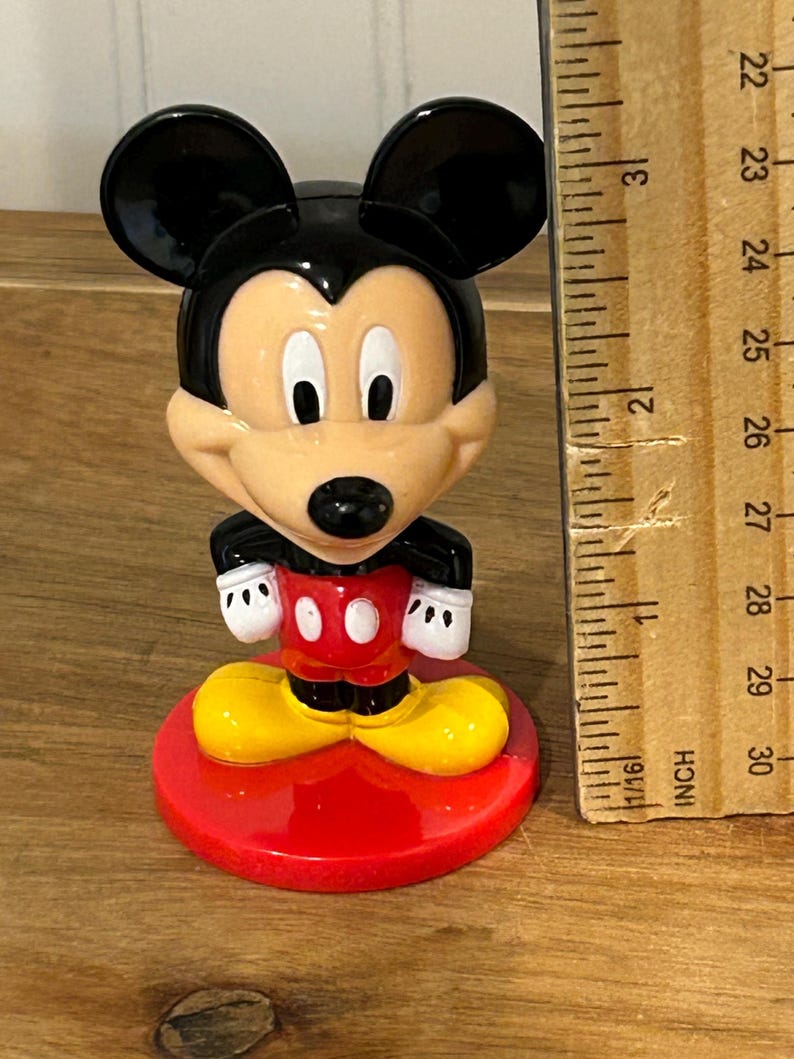 May include: A Mickey Mouse figurine with black ears, a tan face, and a red and white outfit. The figurine is standing on a red base. A wooden ruler is next to the figurine, showing measurements in inches.