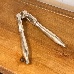 Puede incluir: Un cascanueces plateado con un diseño articulado, apoyado sobre una superficie de madera. El cascanueces está hecho de un metal brillante y tiene un diseño clásico y funcional. Las asas están curvadas para el agarre.