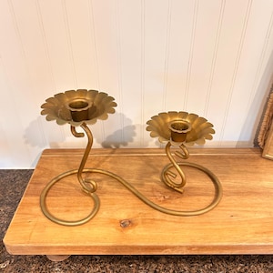 Könnte beinhalten: Ein goldfarbener Metallkerzenhalter mit zwei blütenförmigen Tassen. Der Kerzenhalter hat ein geschwungenes, gewirbeltes Design und steht auf einem Holztablett. Das Gesamtdesign ist elegant und dekorativ.
