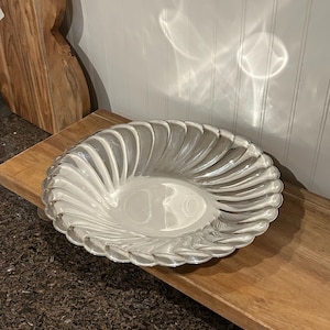 Puede incluir: Cuenco de vidrio transparente con diseño en espiral. El cuenco tiene forma ovalada con borde festoneado. El cuenco está sobre una superficie de madera.