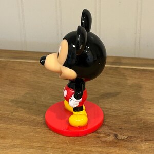 May include: A Mickey Mouse figurine, with a black head and ears, a tan face, and a red, black, and yellow outfit. The figure stands on a red circular base. A classic Disney collectible.