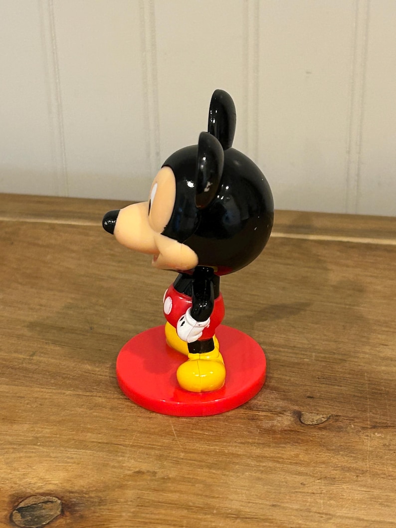 May include: A Mickey Mouse figurine, with a black head and ears, a tan face, and a red, black, and yellow outfit. The figure stands on a red circular base. A classic Disney collectible.