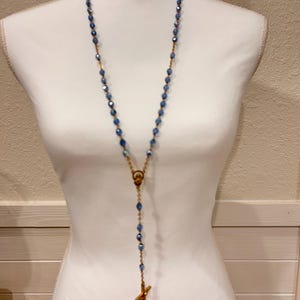 May include: A rosary necklace featuring faceted blue beads strung on a gold-toned metal chain. The necklace includes a central gold-toned connector piece and a gold-toned crucifix pendant hanging at the bottom of the beaded strand.