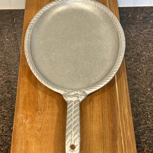 May include: A silver-colored cast iron griddle with a handle. The griddle has a round cooking surface with a rope-like border. The handle features a decorative design and a hole for hanging. The griddle is on a wooden cutting board.