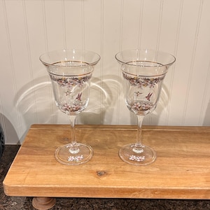 May include: Two clear glass wine glasses featuring a gold rim and a delicate floral pattern with small hummingbirds. The base of each glass is decorated with matching floral motifs, and the stems are slender, leading to a wide, flared bowl shape.