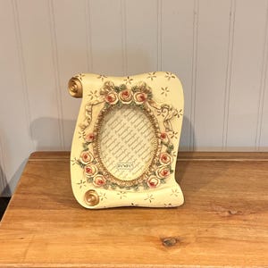 May include: A cream-colored scroll-shaped picture frame featuring an oval center opening surrounded by sculpted pink roses and green leaves. The frame includes a decorative ribbon detail and a label indicating a 2.5 inch by 3.5 inch photo size.