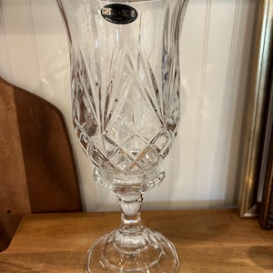 May include: Clear crystal glass candle holder with a flared top and diamond-cut pattern. The candle holder has a pedestal base and a label that reads "Crystal". The glass is transparent, allowing light to pass through.