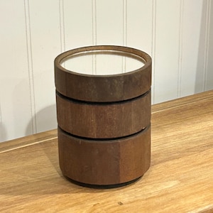 May include: A three-tiered, cylindrical wooden container with a mirrored top. The container is made of dark brown wood and sits on a light brown wooden surface. The background is a white wall with vertical paneling.