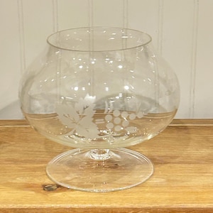 May include: A clear glass brandy snifter with a round bowl and a short stem. The glass is etched with a design of grapes and leaves. The snifter sits on a wooden surface.