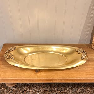 May include: An oval-shaped brass tray with a polished gold-tone finish. The tray features decorative metal accents on both ends, each depicting a cluster of grapes with detailed leaves attached to the rim of the shallow, smooth-bottomed vessel.