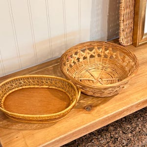 May include: Two woven wicker baskets, one oval and one round, sit on a light brown wooden surface. The oval basket has a solid wooden base, while the round basket has an open weave design. A decorative wicker piece is visible in the background.