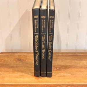 May include: Three black hardcover books by F. Scott Fitzgerald standing upright. The spines feature gold lettering with the titles This Side of Paradise, The Last Tycoon, and The Great Gatsby, along with the author's name and publisher logo at the base.
