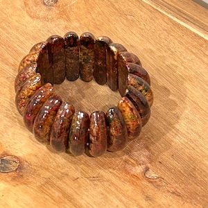 Puede incluir: Una pulsera elástica compuesta por múltiples segmentos rectangulares curvos con un acabado brillante de color marrón oscuro que presenta motas metálicas rojas, verdes y doradas en toda la superficie, creando un aspecto texturizado en cada pieza.
