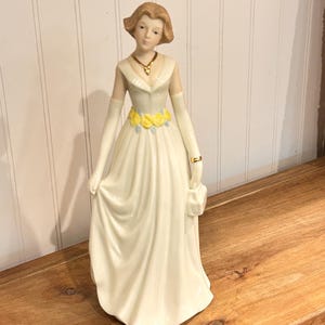 Może przedstawiać: Porcelanowa figurka przedstawiająca osobę z krótkimi brązowymi włosami w długiej kremowej sukni z żółtym kwiatowym detalem w talii. Figurka ma długie kremowe rękawiczki, złoty naszyjnik z wisiorkiem oraz dopasowaną małą torebkę w dłoni.