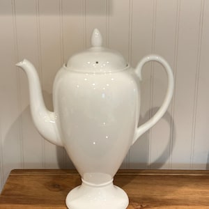 May include: A white ceramic teapot with a lid and a curved handle. The teapot has a spout and sits on a round base. The teapot is on a wooden surface.