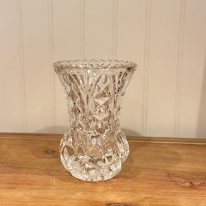 Può includere: Un vaso di vetro trasparente in stile vintage con un motivo a taglio diamante. Il vaso ha una parte superiore svasata e una base arrotondata, che mostra un design classico. Il vetro riflette la luce, evidenziando gli intricati dettagli del cristallo tagliato.