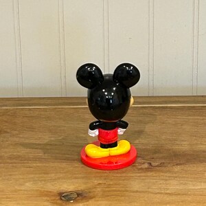 May include: A Mickey Mouse figurine, viewed from the back, stands on a red circular base. The figure has black ears, a black head, a red shirt, yellow shoes, and white gloves. The figurine is on a wooden surface.