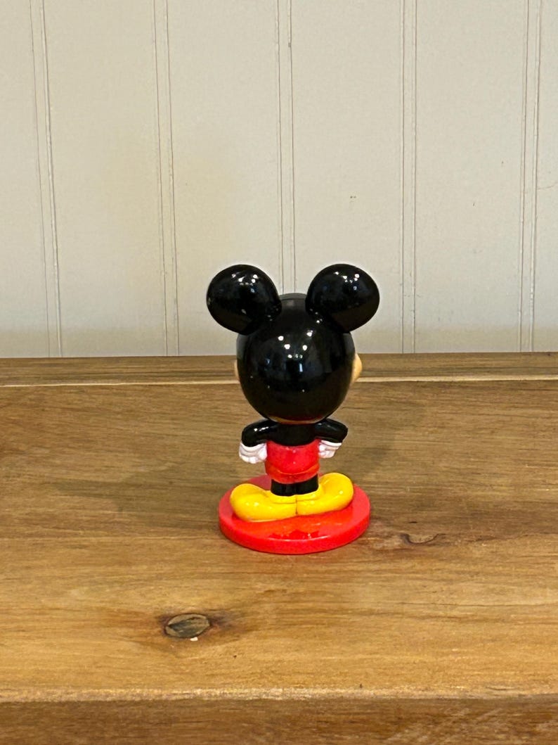 May include: A Mickey Mouse figurine, viewed from the back, stands on a red circular base. The figure has black ears, a black head, a red shirt, yellow shoes, and white gloves. The figurine is on a wooden surface.