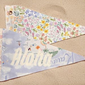May include: Two triangular pennant flags. One features a floral pattern with pink, yellow, and purple flowers. The other has a light blue background with the word "Aloha" in white script and a map design. Both have grommets.