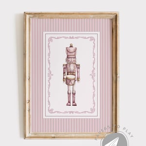 May include: A framed print featuring a watercolor illustration of a pink nutcracker soldier. The artwork is set against a pink striped background and framed in a gold-toned wooden frame. The nutcracker is depicted with a drum and a tall hat.