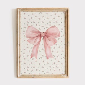 May include: A framed watercolor print featuring a large, soft pink bow. The bow is the focal point, set against a cream-colored background with a delicate floral pattern. The frame has a gold finish, adding a touch of elegance.