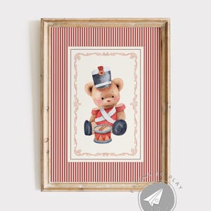 Könnte beinhalten: Ein gerahmter Aquarelldruck eines Teddybären, der als Spielzeugsoldat gekleidet ist. Der Bär trägt eine rote Uniform, einen schwarzen Hut und spielt eine Trommel. Der Rahmen hat ein rot-weiß gestreiftes Muster.