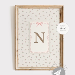 May include: A framed print with the letter "N" in a soft brown colour, set against a cream background with small pink flowers. The print has a decorative pink border and a light, distressed gold frame. Text on the print says "letters A-Z included".