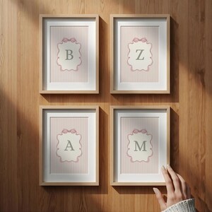 May include: Four framed prints with the letters B, Z, A, and M. Each print features a pink and white striped background with a decorative label and a pink bow. The frames are light brown and mounted on a wooden wall.