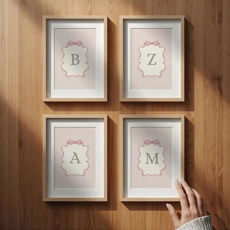 May include: Four framed prints with the letters B, Z, A, and M. Each print features a pink and white striped background with a decorative label and a pink bow. The frames are light brown and mounted on a wooden wall.