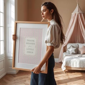 May include: A framed print with the text "THANK HEAVEN for LITTLE GIRLS" in a decorative frame. The print has a pink and white striped background with floral accents. The frame is made of light-colored wood.