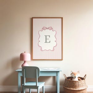 May include: A framed print featuring the letter "E" in a decorative frame with a bow. The print is set against a pink and white striped background. A light blue desk and chair are in front of the print, with a pink lamp and a basket with a stuffed fox.