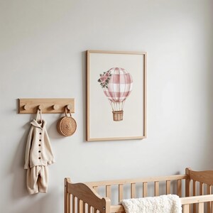 May include: A framed watercolor print of a pink and white checkered hot air balloon with a basket and floral accents. The print is displayed on a light-colored wall, alongside a wooden coat rack with a beige outfit and a woven bag.