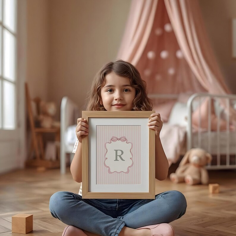May include: A framed print featuring the letter "R" in a light pink and gray design. The print has a light wood frame and is held by a person. The background includes a bed with pink curtains and a stuffed animal.