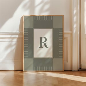 May include: A framed art print featuring the letter "R" in a soft green hue against a neutral background. The artwork is encased in a light wooden frame and has a geometric design in shades of green and beige. The print is displayed against a white wall.