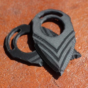 Könnte beinhalten: Zwei schwarze und graue Gitarrenplektren mit Chevron-Design, eines mit rundem Griff. Die Plektren bestehen aus einem strukturierten Material und liegen auf einer rotbraunen Oberfläche.