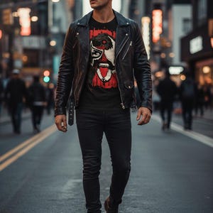 May include: A person wearing a black leather jacket, black graphic t-shirt, black jeans, and black boots. The t-shirt features a red and white graphic design. The person is wearing sunglasses and walking down a city street.