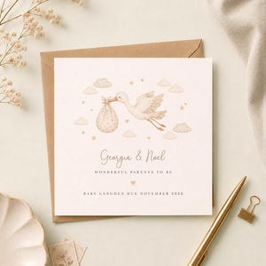 Puede incluir: Una tarjeta de anuncio cuadrada con una ilustración en acuarela de una cigüeña que lleva un hatillo, rodeada de nubes y estrellas. El texto dice: Georgia & Noel, WONDERFUL PARENTS TO BE, y BABY LANGDEN DUE NOVEMBER 2026, con un pequeño corazón.